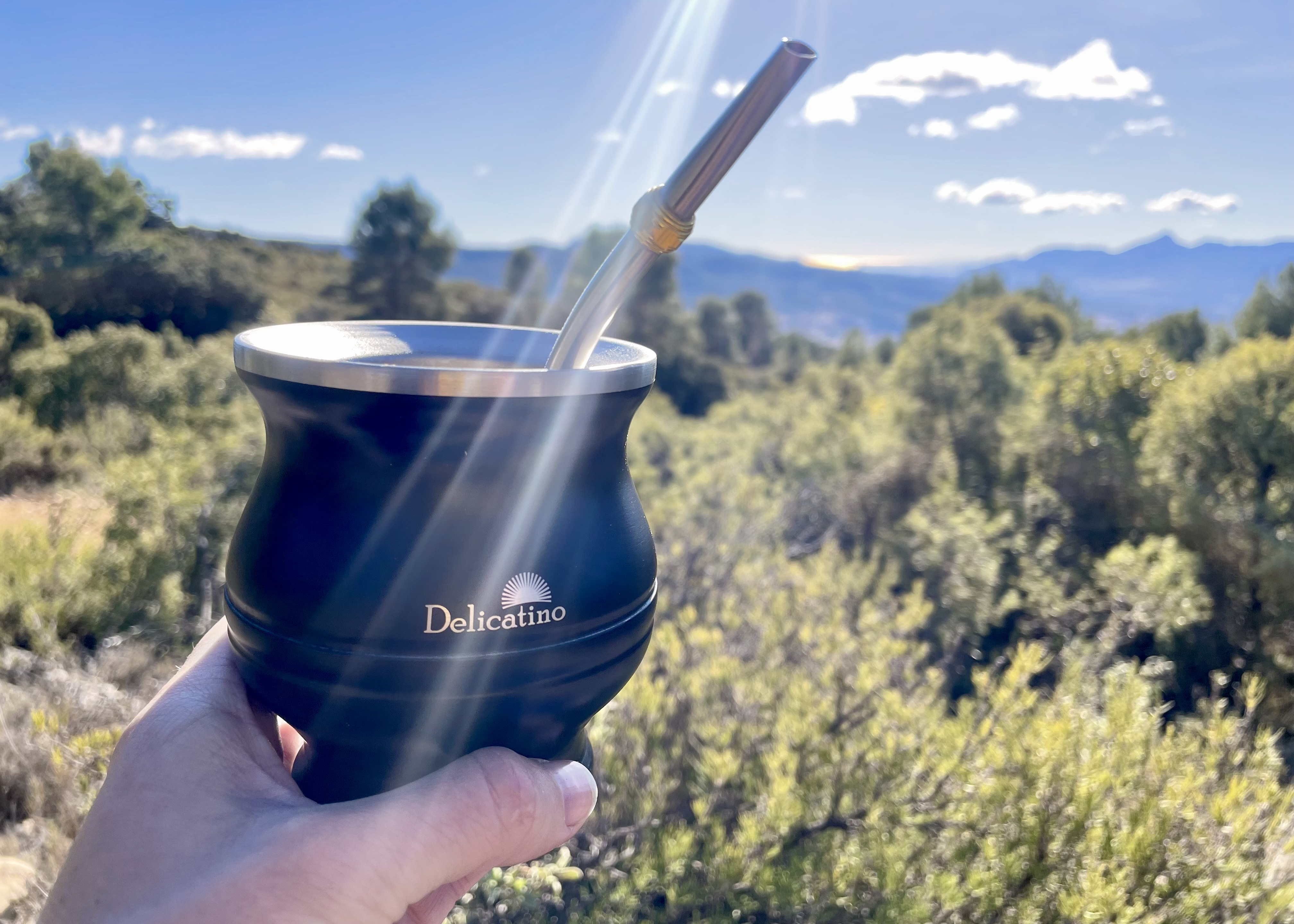 Tomando yerba mate en la montaña, una taza negra (mate) de Delicatino con bombilla con paisaje de montaña de fondo. 