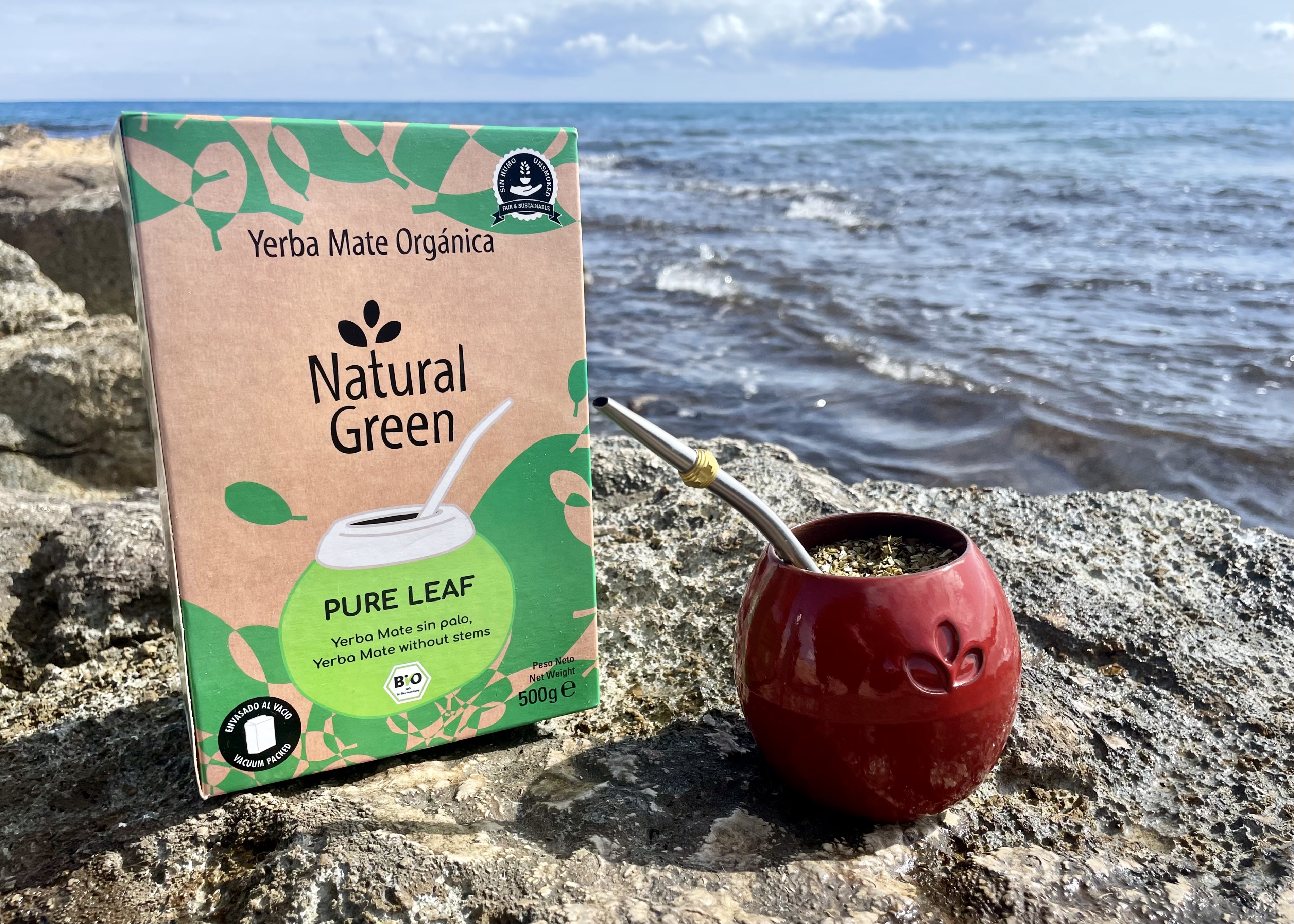 Un paquete de Yerba Natural Green y un vaso de mate rojo sobre una piedra junto al mar.